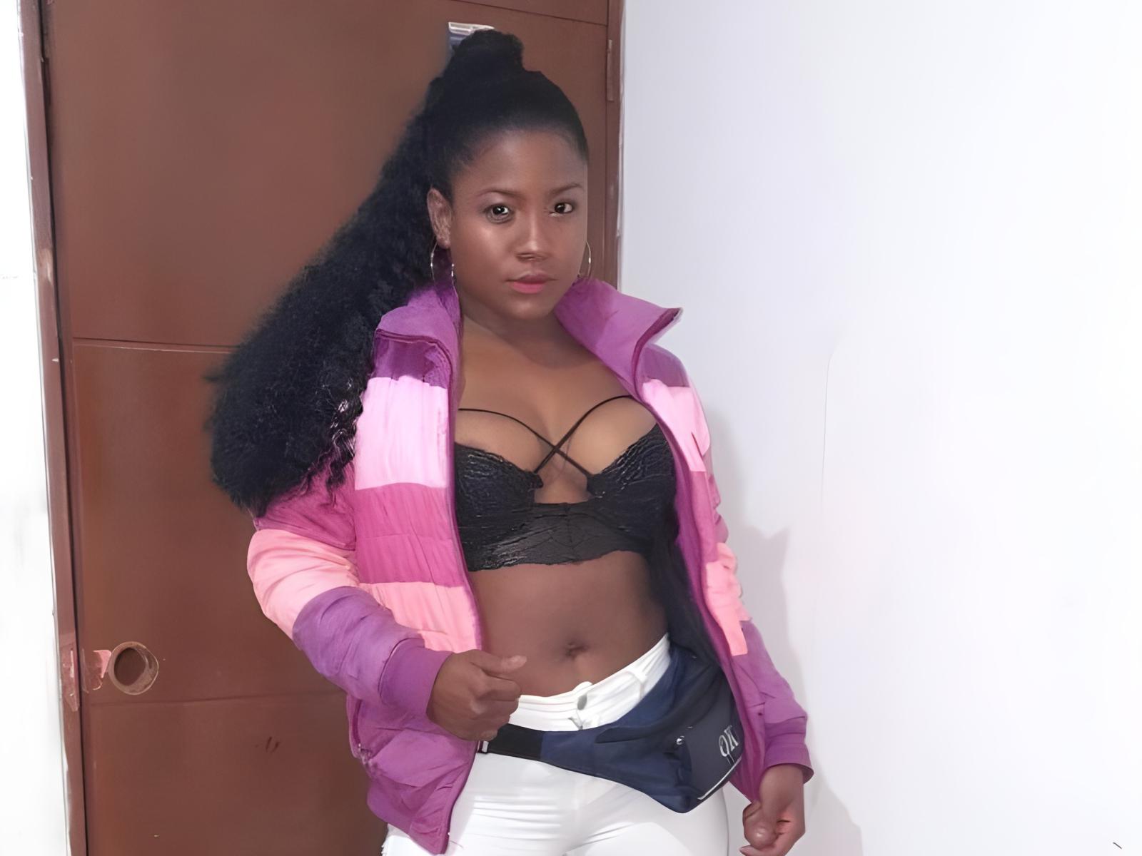 PattyGuerra, com pele escura, cabelo preto encaracolado preso em um rabo de cavalo, vestindo uma jaqueta rosa e roxa, um sutiã de renda preto, calças brancas e um cinto preto, está em frente a uma porta..