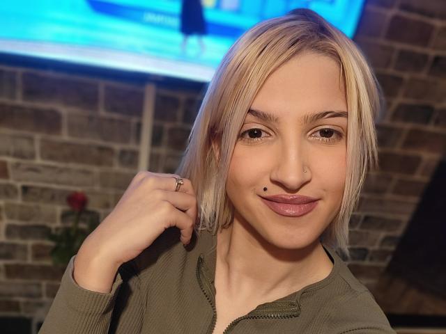 Ajuna with blonde hair, green eyes, and a nose ring, smiling, showing a black mole on her upper lip, wearing a green zip-up, holding her hair.
