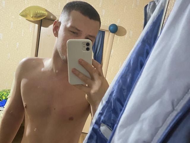 AngelaAndConnie, shirtless with a clean-shaven face, short brown hair, and a small chest, takes a mirror selfie with a white iPhone, showing a blue and white jacket.