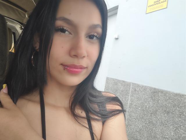 KataleyaMansson with long black hair, pink lipstick, and hoop earrings, smiling slightly, showing a small lip piercing, in a car with gray tiled wall in the background.