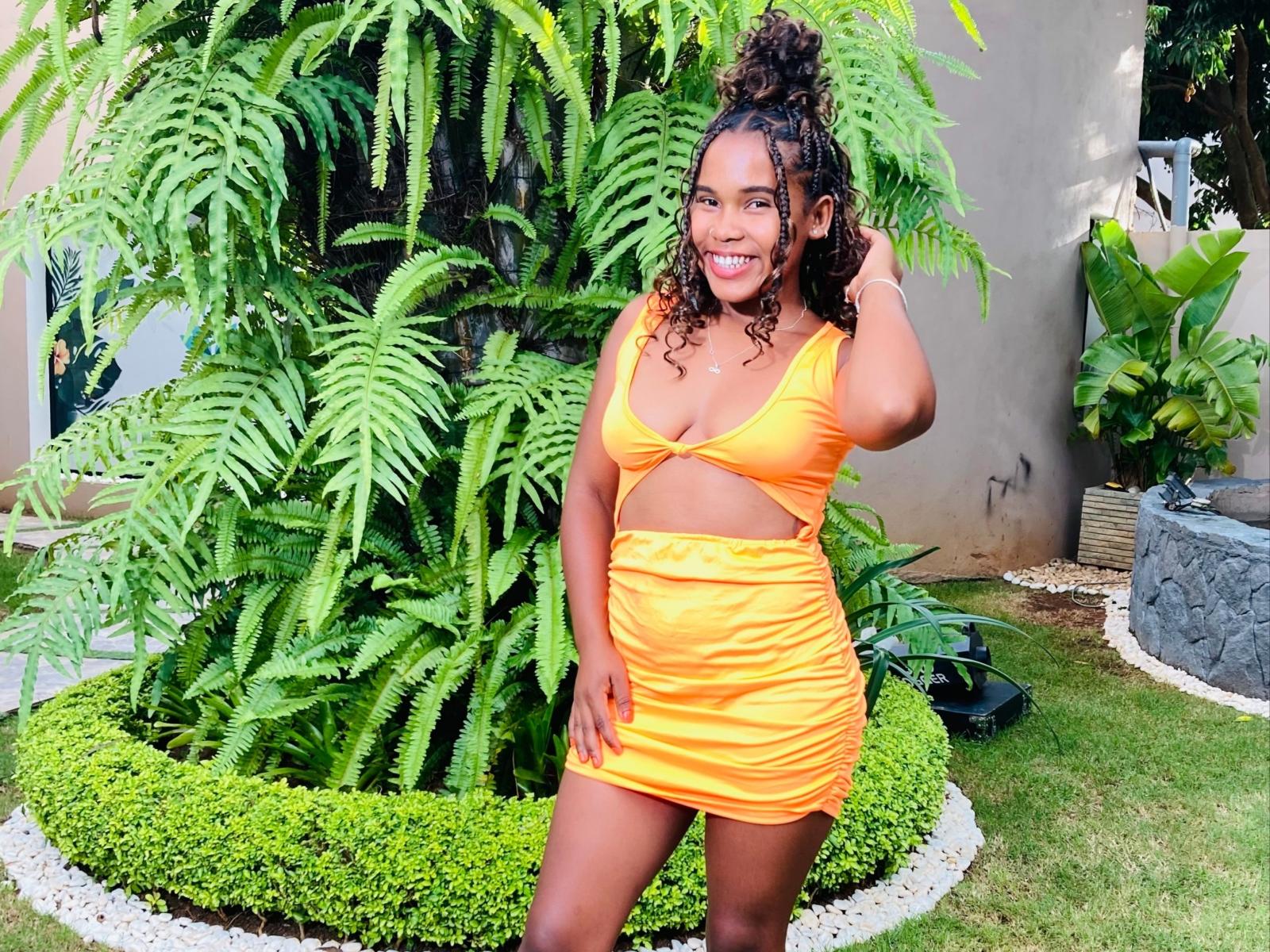 Camilah with curly hair, yellow cutout dress, showing her tits and ass, smiling, hand in hair, standing in front of ferns, green garden, looking sexy and happy.