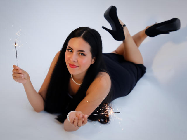 EimmyPark with long black hair, lying on white, holding sparklers in both hands, wearing black dress and high heels, smiling, red lipstick, pearl necklace, ass up in air.