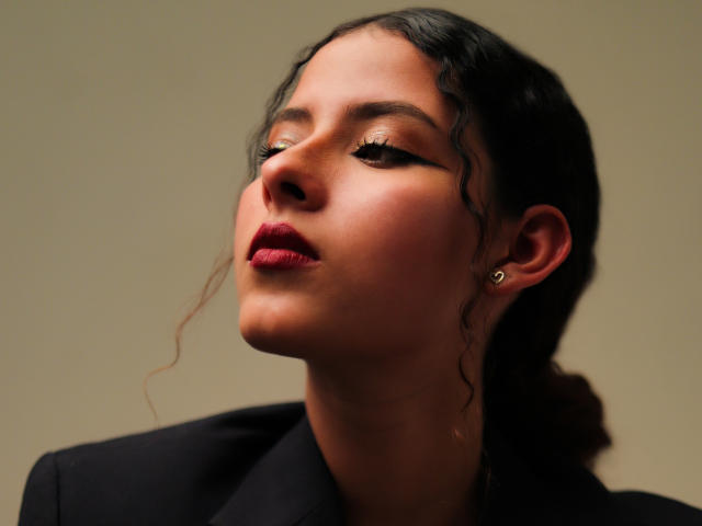 ElenaSantini with wavy black hair, red lipstick, and dark eyeliner, looking up with a slight smile, wearing a black jacket and small stud earrings, against a beige background.
