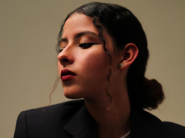 Elena Santini hat welliges, schwarzes Haar zu einem Dutt gebunden, trägt roten Lippenstift und dunklen Eyeliner, eine schwarze Jacke und ein goldenes Ohrring. Ihre Augen sind geschlossen und ihr Gesichtsausdruck ist ernst..