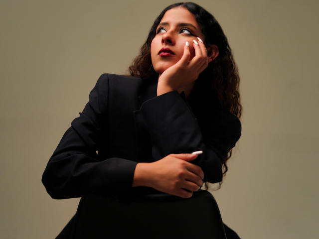 Elena Santini, con i capelli ricci e scuri, indossa un tailleur nero, con una mano sul viso, che guarda verso l'alto, rossetto rosso, con la testa leggermente inclinata, con un'espressione pensierosa, e un braccio incrociato..
