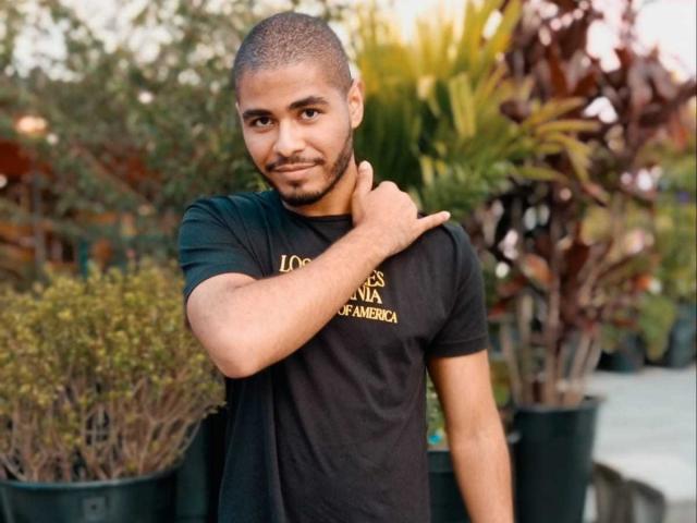 JhonOud with short black hair, dark skin, and a beard, wearing a black t-shirt, touching his neck, standing in front of potted plants, smiling slightly.