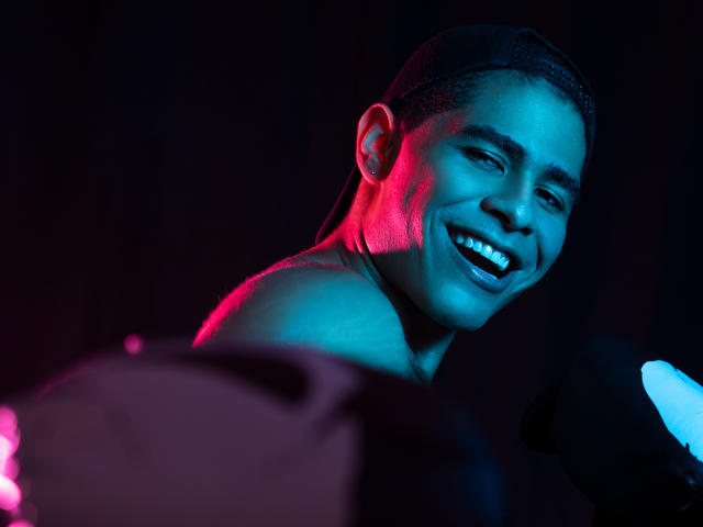 JhonOud, smiling with a toothy grin, wearing a backwards cap, blue and pink lighting on his dark skin, shirtless, holding a microphone in a dimly lit setting.