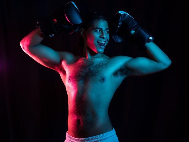 JhonOud, shirtless with black boxing gloves, has a muscular chest with hairy nipples, white pants, and a happy grin, illuminated by pink and blue lights.
