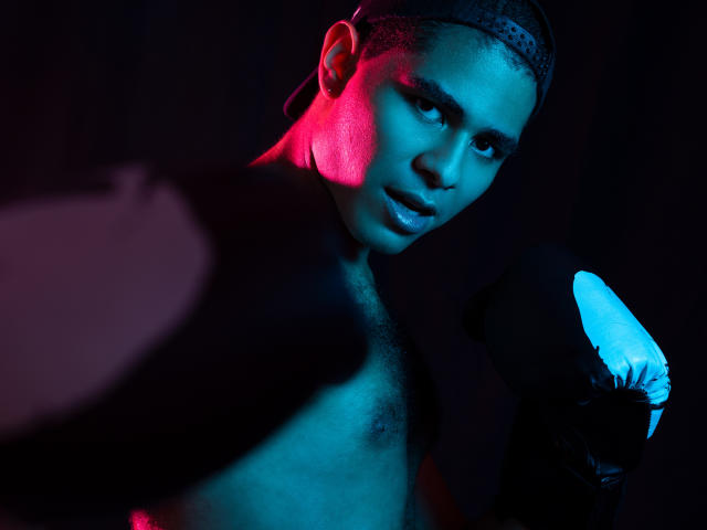 JhonOud with dark skin, short hair, and a light beard, wearing black gloves, under blue and pink lighting, looking at the camera with a serious expression.