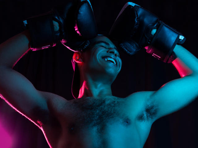 JhonOud, shirtless with black boxing gloves, flexing muscles, smiling with eyes closed, showing hairy chest and armpits, lit with pink and blue lights.