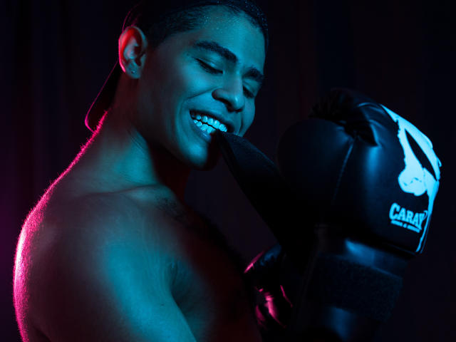JhonOud, shirtless, holding black boxing gloves, smiling, blue and pink lights, dark background, sweat on face, short hair, muscular, right hand near mouth, gloves marked "CARANO".