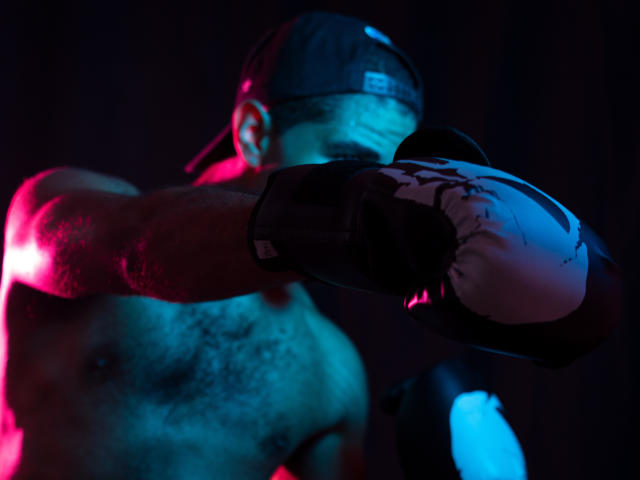 JhonOud, shirtless with a hairy chest, wearing a dark baseball cap and black boxing gloves, punches forward in a blue and pink-lit image.