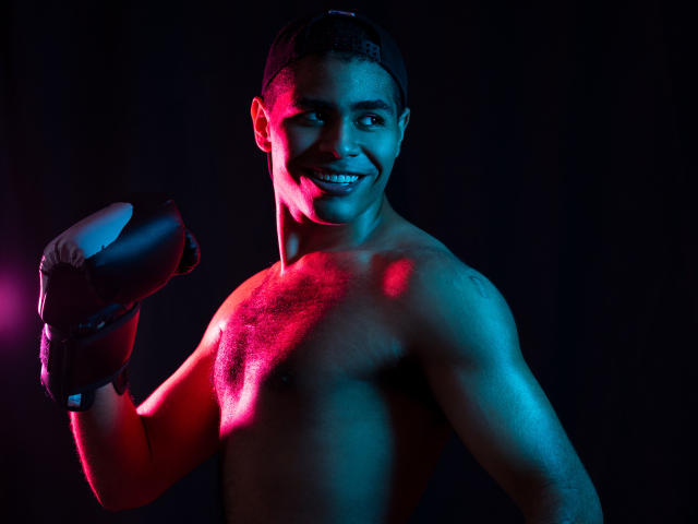 JhonOud, shirtless, with black boxing gloves, raised fist, wearing a backward cap, blue and pink lighting, smiling, showing a toned, muscular chest and a confident, sexy look.