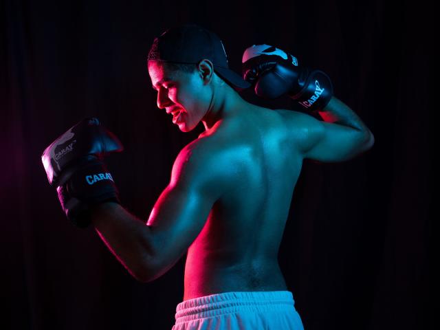JhonOud, shirtless with black gloves labeled "CARA," white pants, and backward cap, stands in profile, muscles glistening under blue and pink lights, hands up in boxing stance.