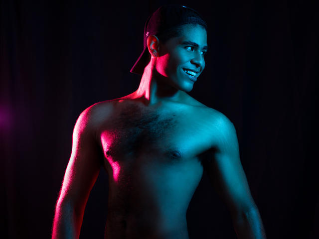 JhonOud, shirtless, with a black backward cap, dark skin, and a handsome smile, showing a muscular chest with small, dark nipples and a subtle chest hair. Blue and pink lights.