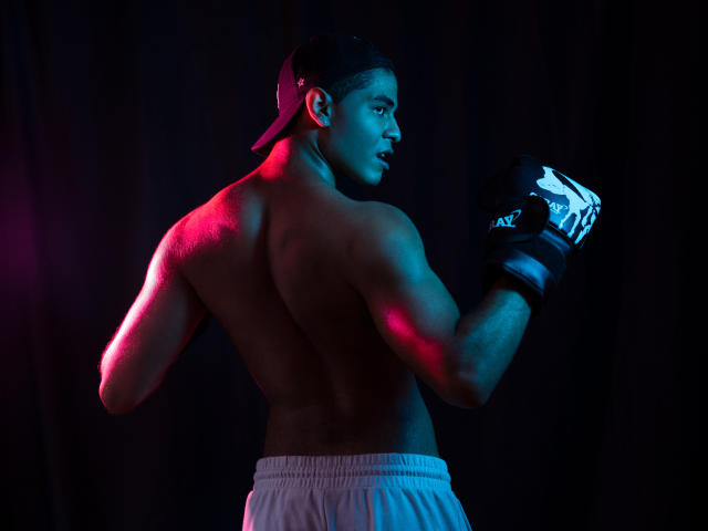 JhonOud is shirtless, muscular, with a backward baseball cap, white shorts, and black boxing gloves, standing in a dark room with red and blue lighting.