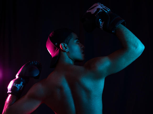 JhonOud's shirtless, muscular back with arms raised, wearing black boxing gloves, and a backward cap, lit by blue and pink lights, showing defined arms and strong posture.