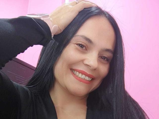 CharlotteCrofort with long black hair, red lipstick, and a nose piercing, smiling while a hand with pink gloves touches her head. She's wearing a black top.