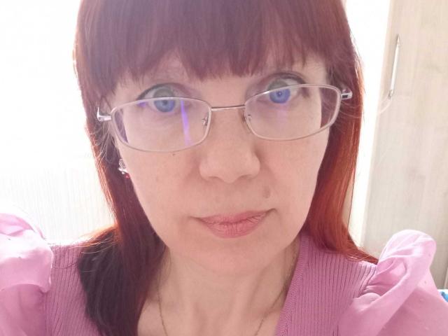 JuicyLady69 has red hair, glasses, and pink puffed-sleeve shirt. She has pale skin, blue eyes, and a small beauty mark on her right cheek. Her lips are slightly puckered.