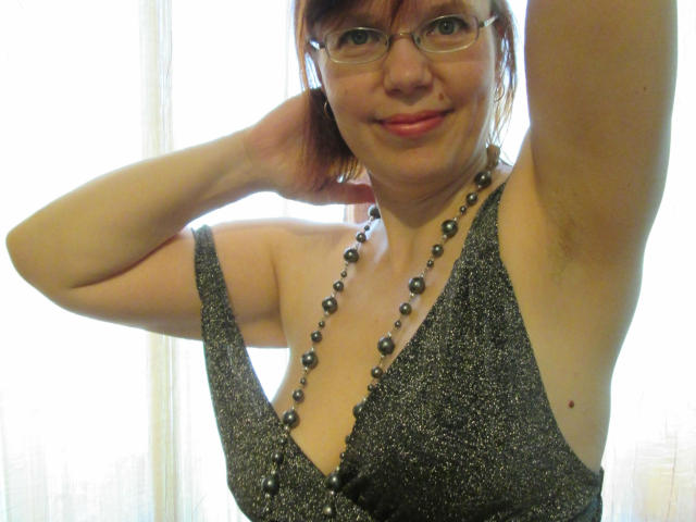 JuicyLady69 with glasses, brown hair, smiling, wearing a glittery gray dress with a deep V-neck, black bead necklace, and her arm raised, showing a slightly arched eyebrow.