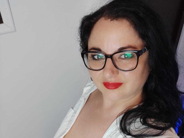 AleshaArabian has green eyes, black glasses, red lipstick, and wavy black hair. She's wearing a white shirt, showing her tits slightly. She's smiling subtly.