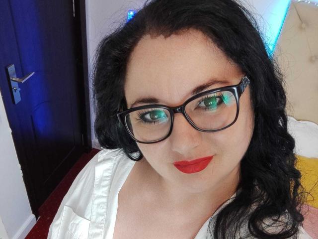 AleshaArabian with glasses, red lipstick, and black wavy hair, wearing a white shirt, has green reflective eyes and a slight smile, standing near a dark door.