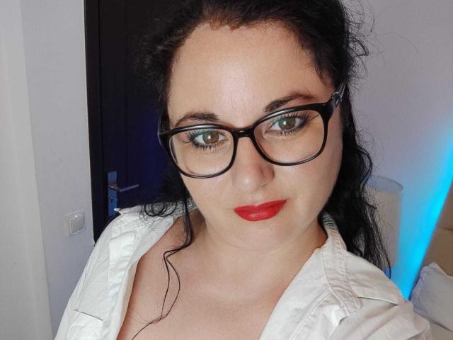 AleshaArabian with glasses, red lips, white shirt, and black hair, showing green eyes, slightly chubby face, and a serious expression. Boobs partially visible under shirt.