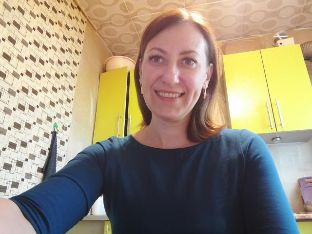 AshleyXHoney with shoulder-length brown hair, blue eyes, and a smile, wearing a navy blue shirt, standing in a kitchen with yellow cabinets and patterned walls.