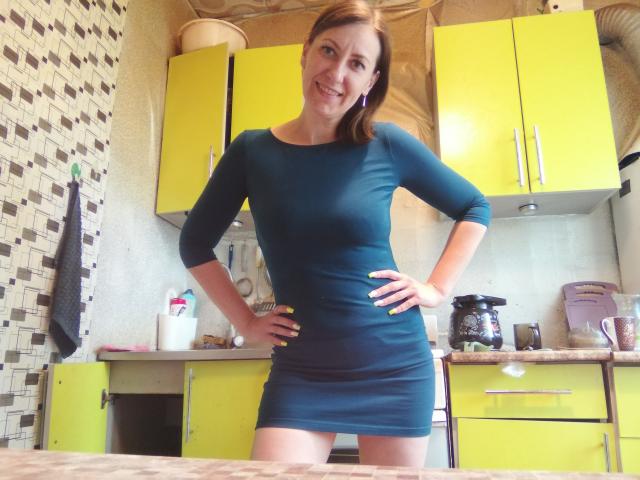 AshleyXHoney se tient dans une cuisine, vêtue d'une robe turquoise moulante, les mains sur les hanches, souriante, avec des fesses apparentes et des seins de taille moyenne, des armoires jaunes et une machine à café en arrière-plan..