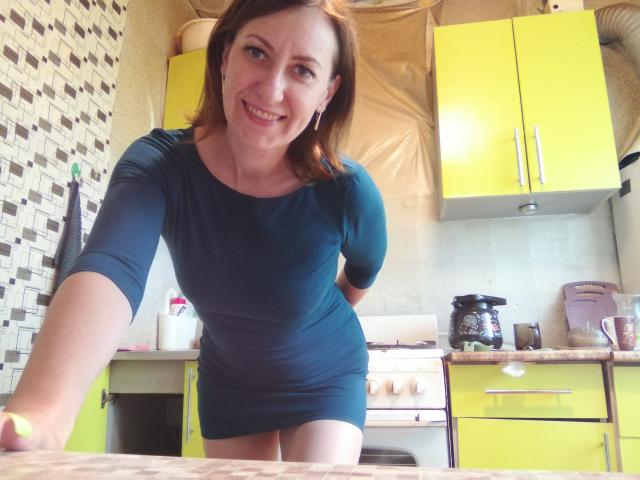 AshleyXHoney mit mittlerer Brustgröße, trägt ein eng anliegendes, türkisfarbenes Kleid, lehnt sich vor und lächelt in die Kamera, in einer gelben Küche mit gelben Schränken und einem weißen Herd..