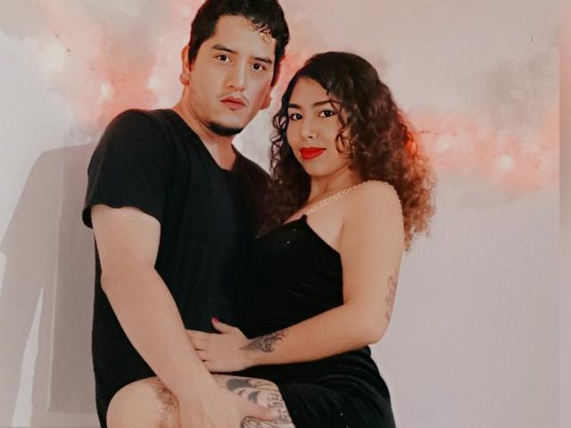 SamyAndZack, com cabelo cacheado, batom vermelho, vestido preto e tatuagens, sentada no colo de SamyAndZack, vestindo uma camisa preta, mostrando pernas peludas, segurando as ancas uma da outra..