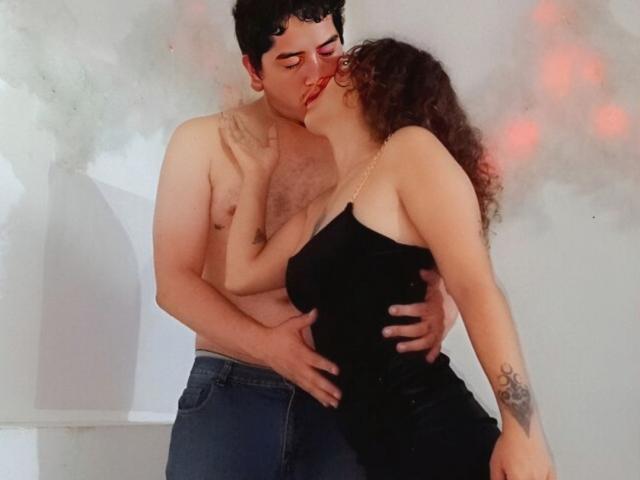 SamyAndZack, senza maglietta e con una corporatura robusta, bacia Zack, che ha i capelli ricci e indossa un abito nero, mentre si tiene le natiche. La mano di Zack è sui seni di Zack, che ha un tatuaggio sul braccio destro..