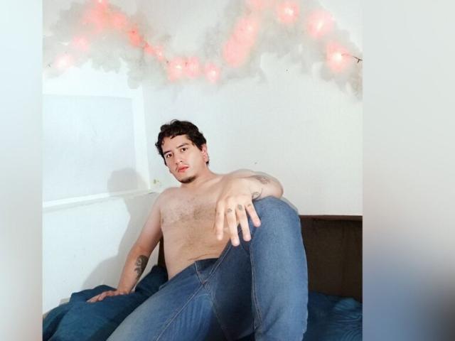 Samy e Zack senza camicia, con capelli ricci scuri, jeans e un tatuaggio sul braccio sinistro, seduti su un letto con un'espressione rilassata, leggermente seducente..