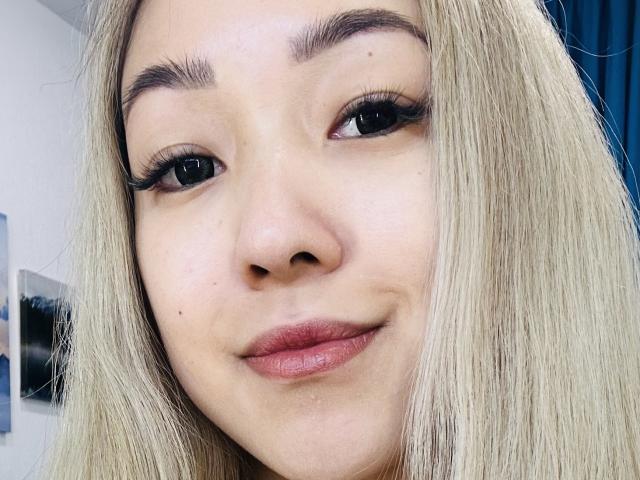 RenyLime's face with blonde hair, thick eyebrows, long eyelashes, and pink lips, slightly smiling, showing a small mole on her left cheek. Her eyes are looking directly at the camera.