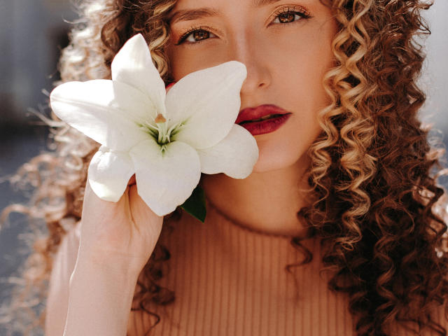 BiancaHotLove with curly brown hair holding a white lily over her face, wearing a ribbed turtleneck, has red lipstick and brown eyes, looking sexy.