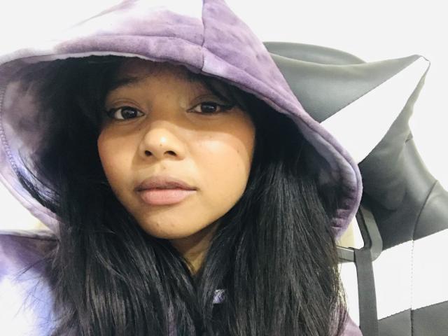 Omaliia, con piel de tono marrón medio, cabello negro y lacio, vistiendo una capucha morada con forma de gato, con una expresión ligeramente aburrida y los labios ligeramente entreabiertos, sentada en una silla blanco y negra..
