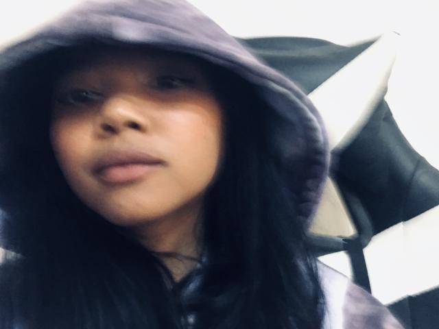 Omaliia, avec ses cheveux foncés et portant une capuche violette, a une expression neutre et est assise sur un fauteuil de jeu noir et blanc. Son visage est flou, sa peau est légèrement brillante. L'éclairage est doux..