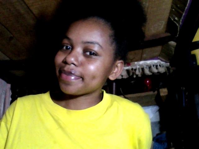 RayOfBeauty with dark skin, black curly hair, and shiny lips, wearing a yellow shirt, smiling slightly, looking at the camera.