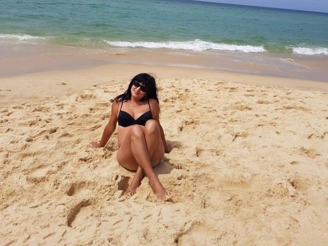 SweetMonyk with black bikini, sitting on sandy beach, legs crossed, arms resting, black hair, sunglasses, looking up, ocean and waves in background, sunny day, tan skin.