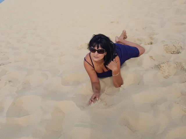 SweetMonyk with black hair, wearing sunglasses and a blue dress, on all fours in sandy beach, her ass slightly raised, showing a tan line on her back.