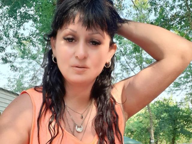 SweetMonyk has wavy black hair, brown eyes, and wears a pink sleeveless top with silver necklaces. She's touching her head, showing off small hoop earrings.