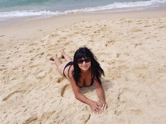 SweetMonyk with black hair, sunglasses, and a black bikini, lying face-down on sandy beach, her tits and ass slightly visible, smiling, legs bent at knees, arms in front.