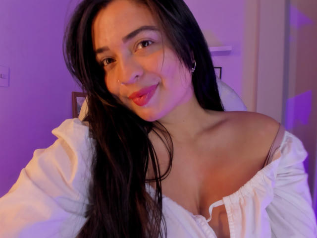 KylieFire with long black hair, smiling, wearing a white off-shoulder top, exposing her small tits, pink lipstick, and small hoop earrings, under purple lighting.