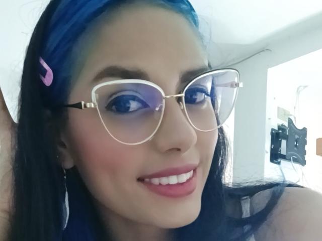 Anais Hilton mit blauen Haaren, einer rosa Haarspange und einer klaren Brille, lächelnd mit pinkem Lippenstift, wobei weiße Zähne zu sehen sind, und mit einem süßen Gesicht, in einem hellen Innenraum..