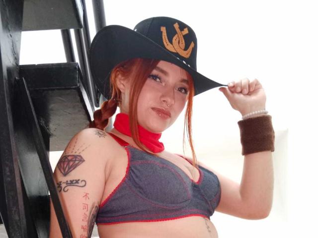 Antokira with red braids, wearing a black cowboy hat, gray sports bra, red scarf, brown wristband, tattoos, nose ring, standing by black stairs, holding hat.
