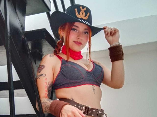Antokira with red bandana, black cowboy hat, and tattoos, wearing a gray bra, brown wristbands, and belt, standing next to a black staircase, showing her small tits.