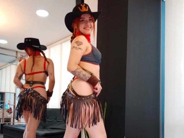 Antokira with red hair, cowboy hat, gray bra, and brown fringed skirt, showing tattoos, smiling, standing, reflecting in mirror, revealing butt, small tits, and red bra straps.