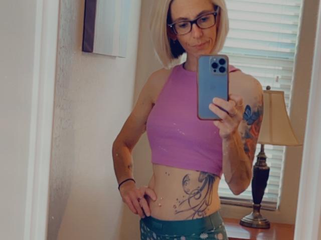 IttyBitty with blonde bob, glasses, purple sports bra, green pants, tattoo on right side, holding phone, smiling, standing in front of mirror, showing belly button.