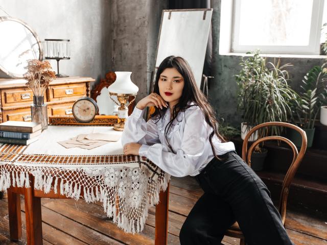 AnneTLuis with long dark hair, white button-up shirt, black pants, leaning on a wooden table with a white lace cloth, red lipstick, looking at the camera, indoors.