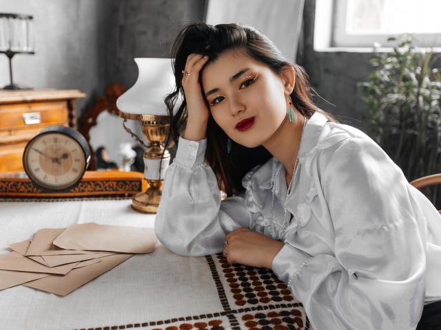 AnneTLuis, Asian woman, white ruffled blouse, red lipstick, black hair, hand on head, lying on table, lamp, clock, envelopes, patterned cloth, looking at camera, soft smile.